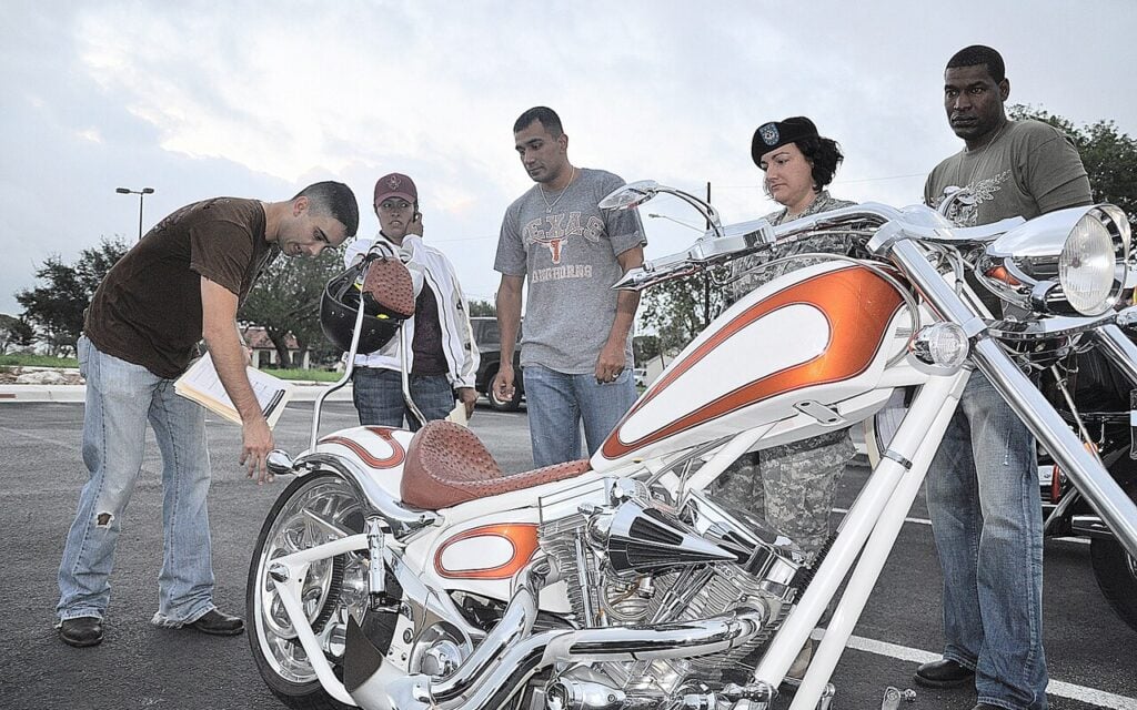 Des acheteurs inspectent une moto d'occasion, papiers en main, pour contrôler les documents et l'état avant l'achat » Légende : Photo : Esther Garcia / U.S. Army, domaine public, via Wikimedia Commons