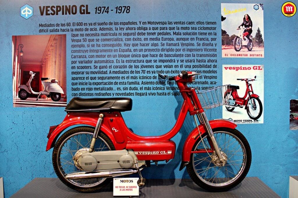 Un Vespino GL rouge de 1983 exposé au musée devant des publicités d'époque