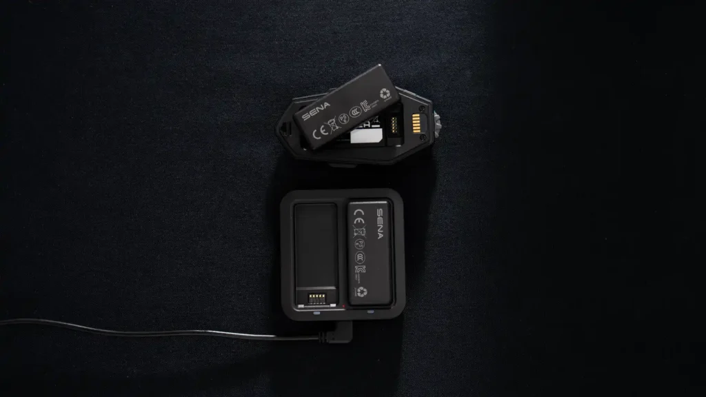 Intercom Sena Apex ouvert avec sa batterie amovible retirée, posé à côté du chargeur double fourni (Source : Sena)